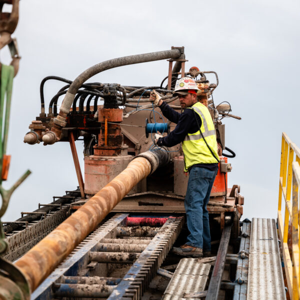 Worker in neon vest guides pipe with horizontal directional drill at utility construction site.