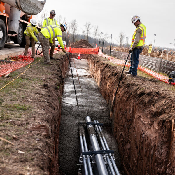 Construction workers in safety gear supervise concrete pouring over high-voltage conduit pipes.