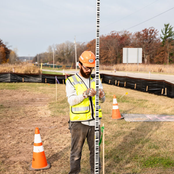 Construction worker measures roadside with survey rod near cones.