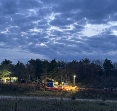 Construction site with machines, bright lights, trees, cloudy dusk.