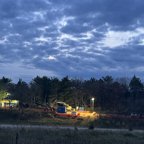 Construction site with machines, bright lights, trees, cloudy dusk.