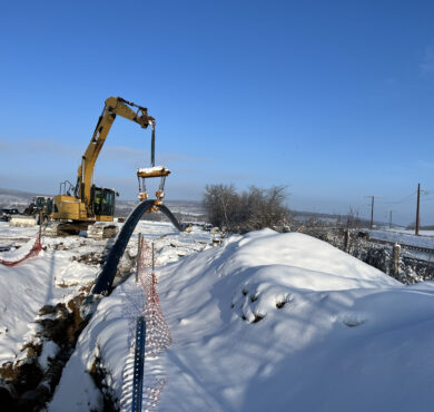 Excavator lifts pipe over snowy trench as workers oversee.